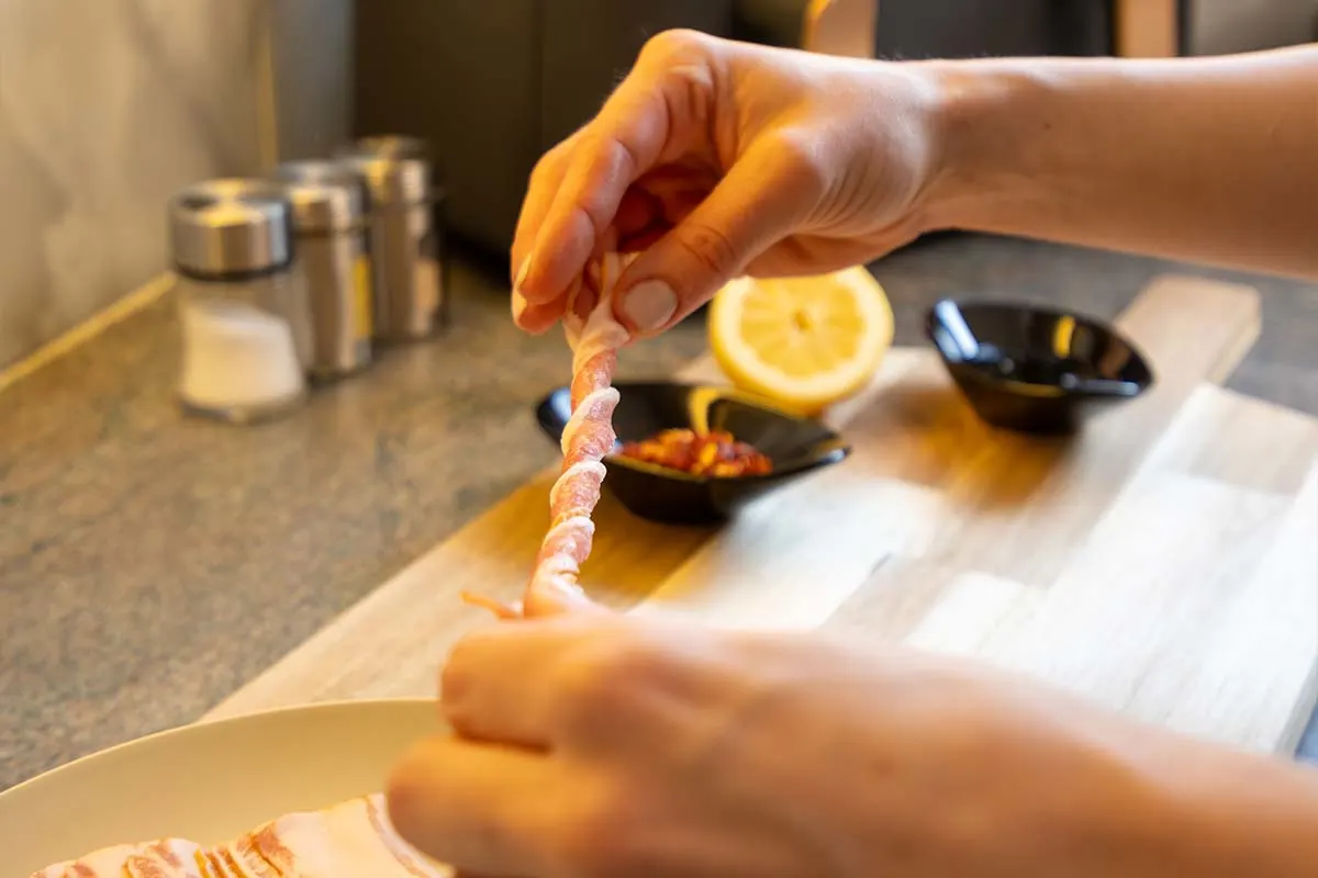 Hände wickeln Speckstreifen um ein Stück Fingerfood auf einem Schneidebrett in der Küche. 