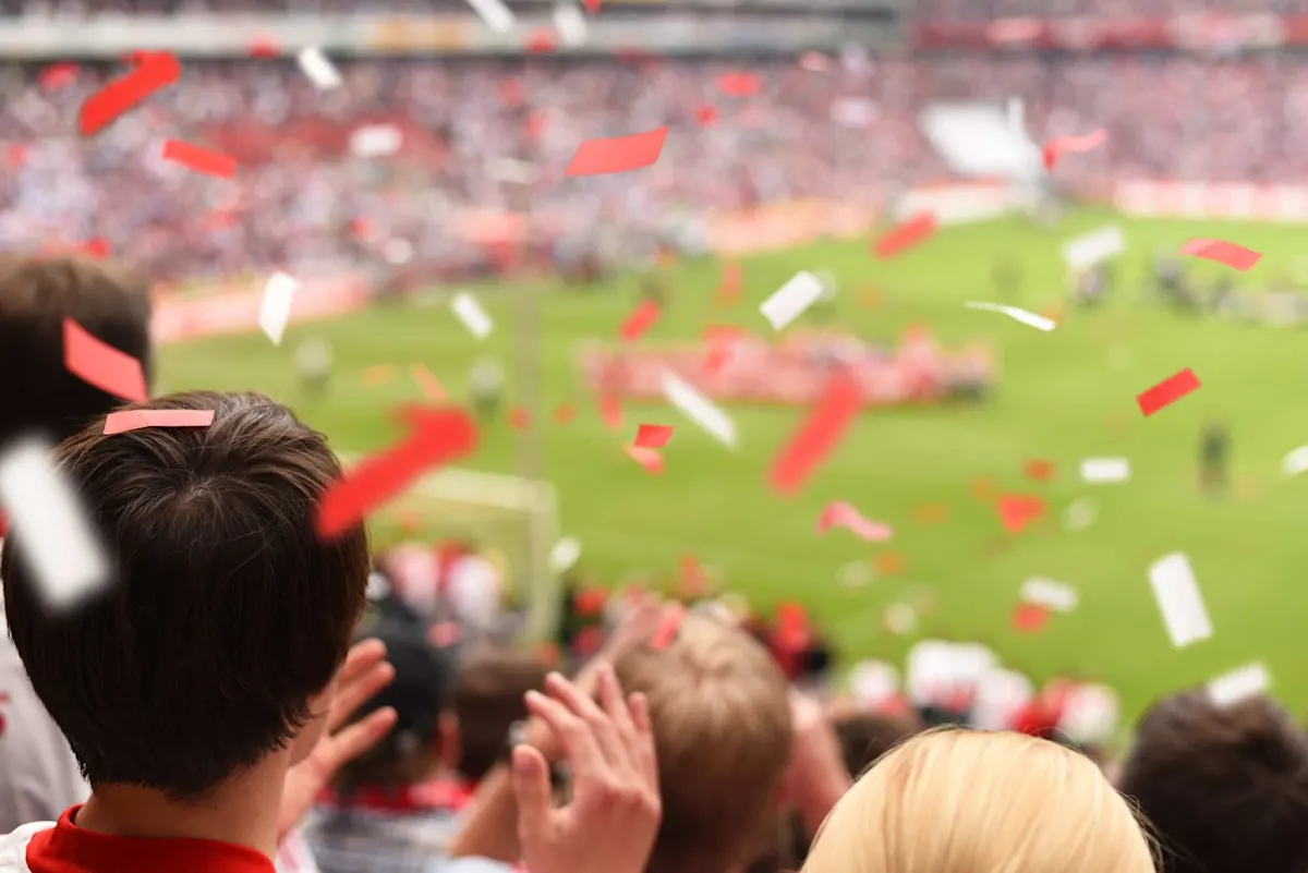 Fussballfans stehen im Stadion und jubeln, während rot-weisses Konfetti durch die Luft fliegt.