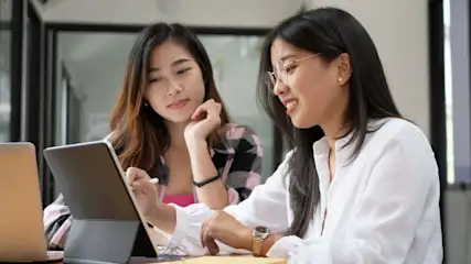 Due donne lavorano insieme su un tablet, accanto a un computer portatile e a dei quaderni.