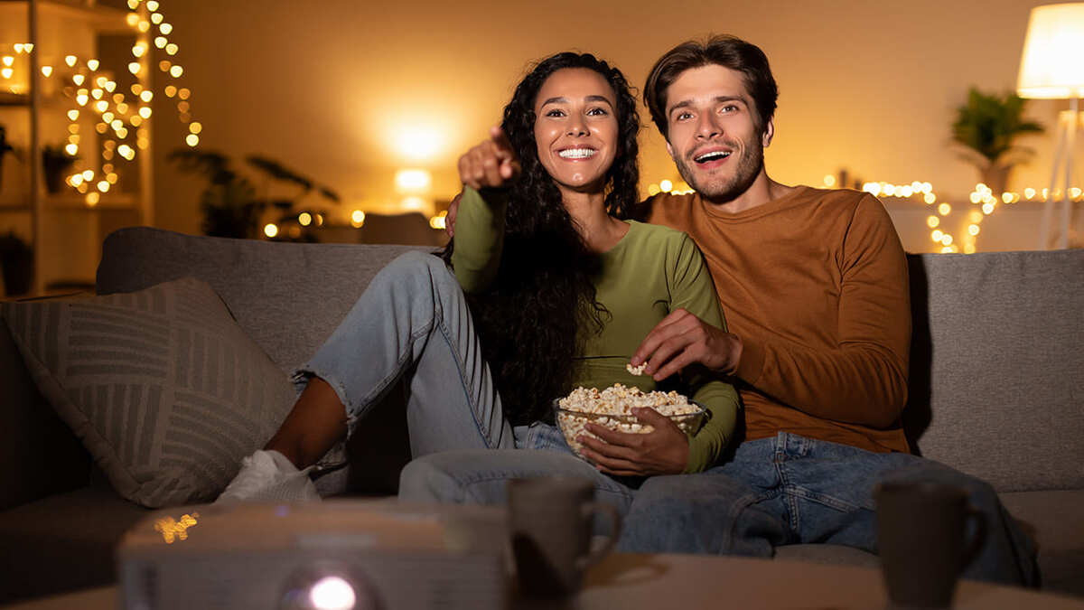 Un couple regarde un film avec un projecteur, assis sur un canapé avec du pop-corn. Guirlandes lumineuses décoratives en arrière-plan.