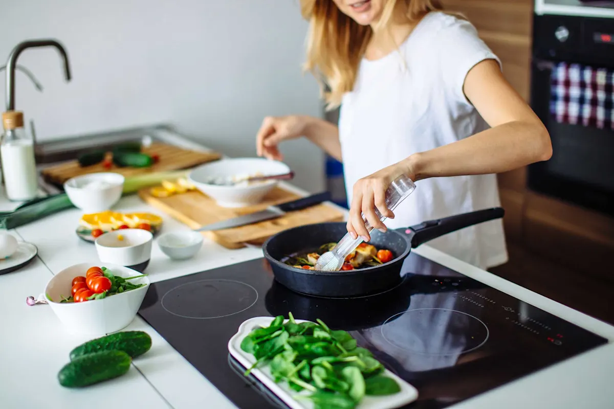 Femme dans la cuisine, manipule une poêle avec une pince, d'autres ustensiles de cuisine à côté.