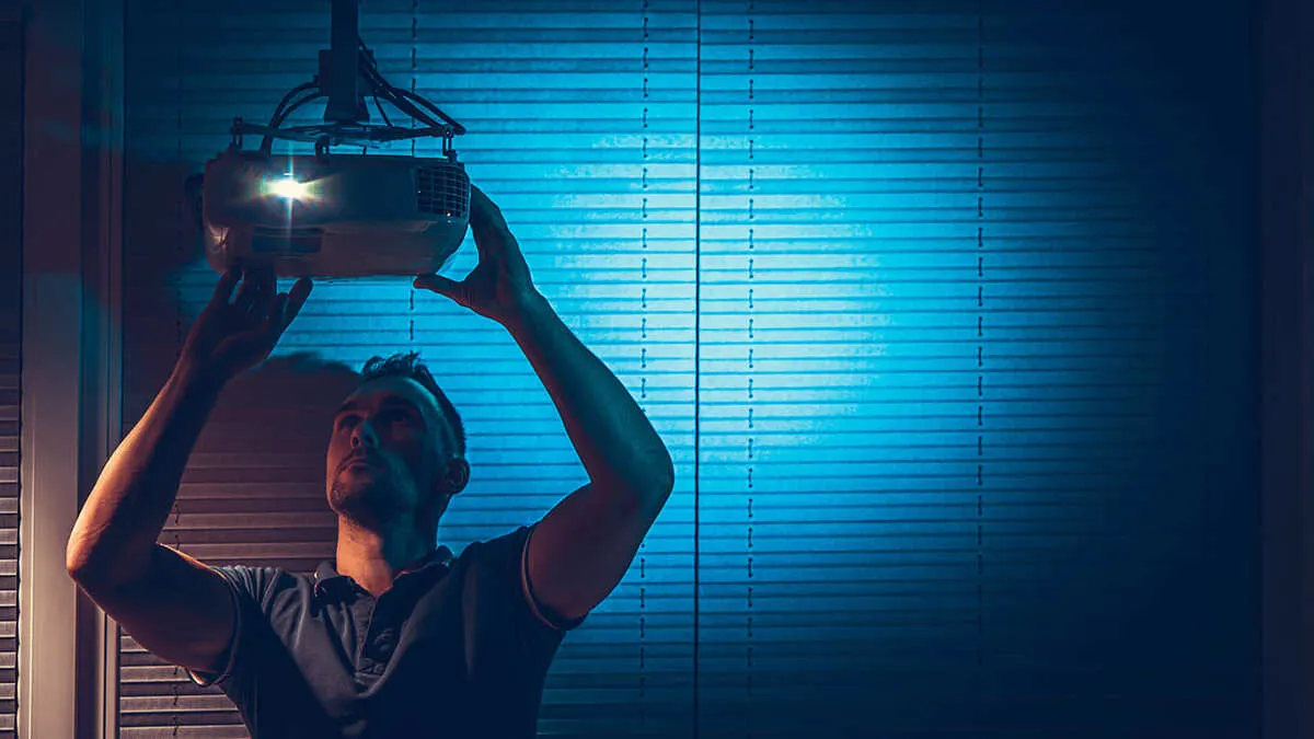 Ein Mann montiert einen Projektor an der Decke vor einem blauen Rollo. Das Licht des Projektors scheint hell.