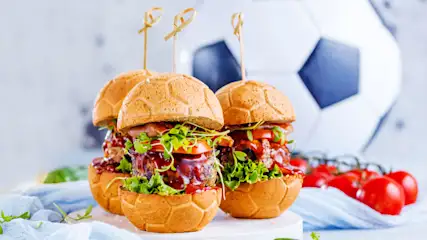 Drei Fussball-Burger mit Salat, Tomaten und Fleischfüllung, mit Zahnstochern befestigt, vor einem Fussball und Tomaten.