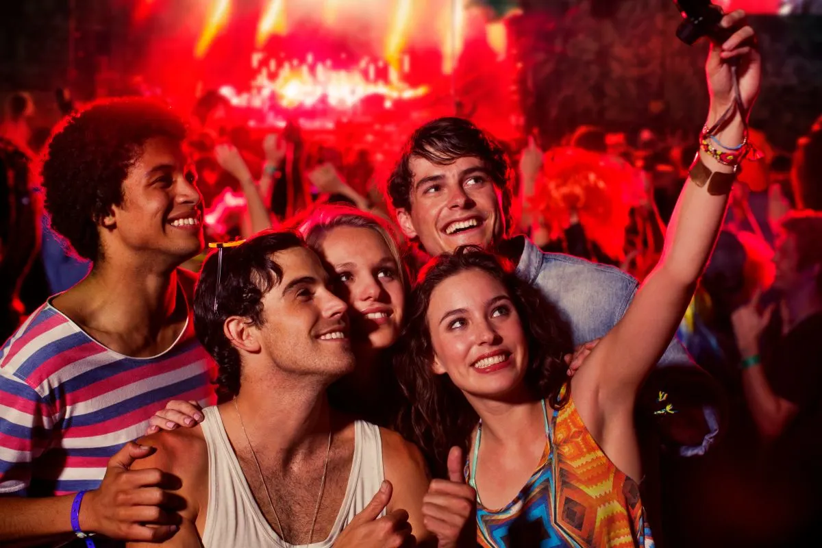 Gruppe junger Leute macht ein Selfie auf einem Openair-Festival, im Hintergrund spielt eine Band auf der Bühne.