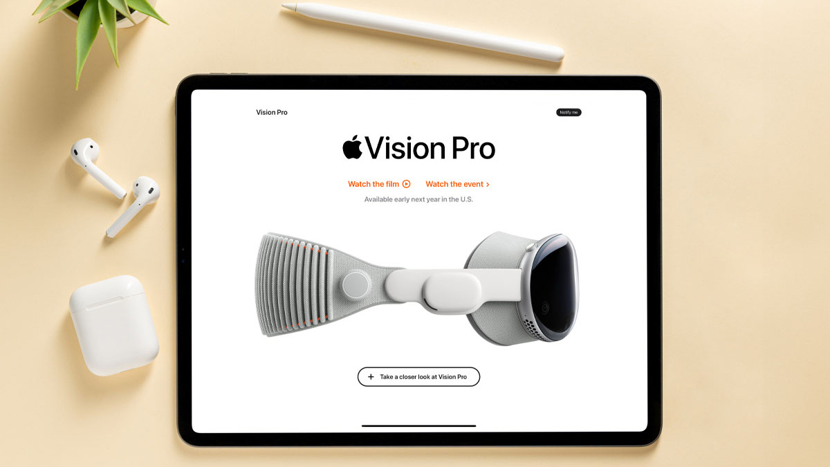 Auf einem Tisch liegt ein Tablet mit der Vision Pro-Website, daneben Kopfhörer, ein Stift und eine Topfpflanze.