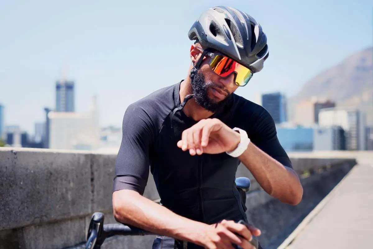 Mann mit Vollbart, Fahrradhelm und verspiegelter Sportbrille schaut während einer Radtour in der Stadt auf seine Smartwatch, im Hintergrund unscharfe Skyline.