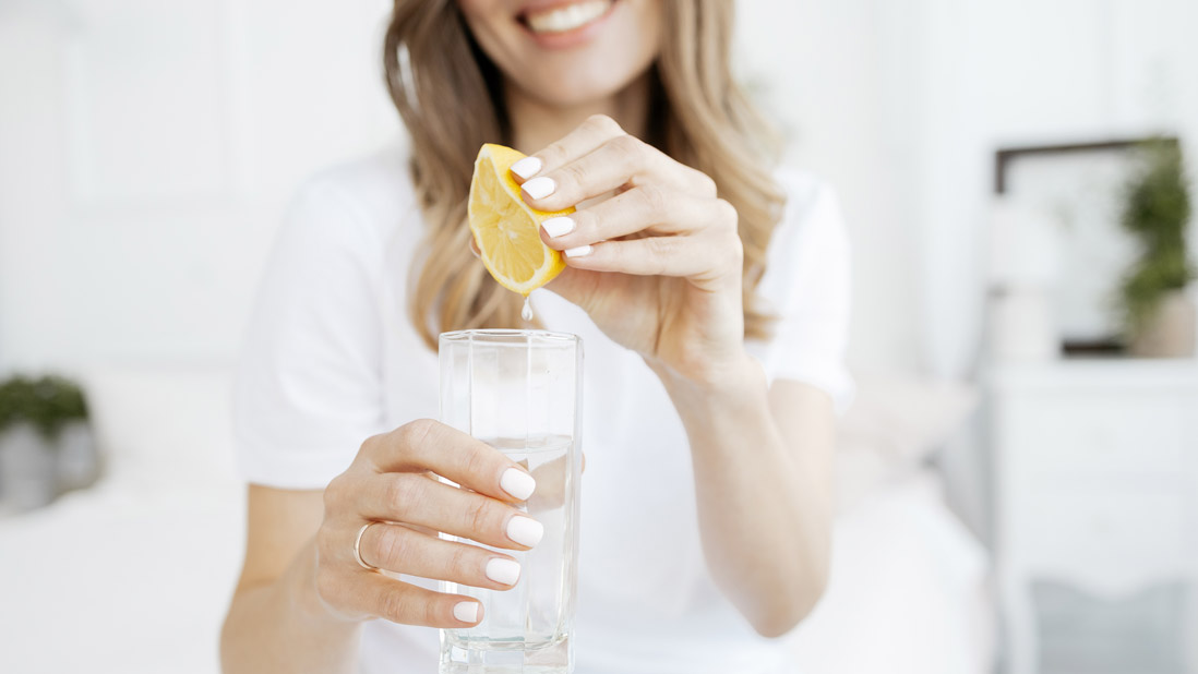 Une femme presse un citron jaune dans un verre d'eau. L'arrière-plan est flou et comprend un cadre et une plante.