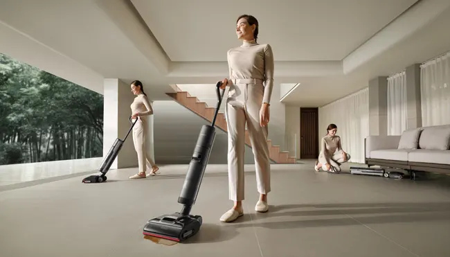 Une femme passe l'aspirateur dans une pièce lumineuse et aérée avec des fenêtres, un escalier et un canapé.