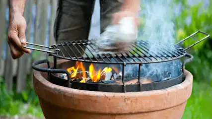 Un gril en métal au-dessus d'un foyer en argile contient du charbon et des flammes, produisant de la fumée, sur fond de verdure.