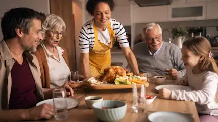 Una donna con il grembiule serve a una famiglia seduta a tavola un pollo arrosto intero.
