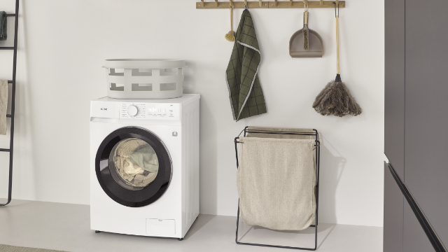 Λευκό KOENIC KWDR 8642 Πλυντήριο-στεγνωτήριο INV σε μοντέρνο δωμάτιο πλυντηρίων, δίπλα του ένα καλάθι ρούχων και αξεσουάρ.