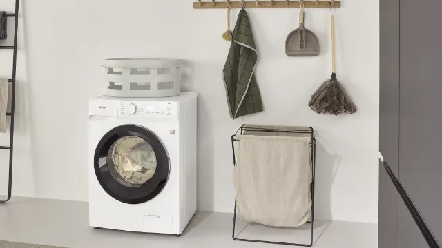 Λευκό KOENIC KWDR 8642 Πλυντήριο-στεγνωτήριο INV σε μοντέρνο δωμάτιο πλυντηρίων, δίπλα του ένα καλάθι ρούχων και αξεσουάρ.