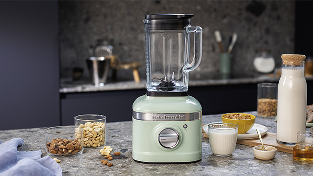Ein hellgrüner Standmixer steht auf einer Küchentheke, umgeben von Zutaten wie Nüssen, Milch und Honig.