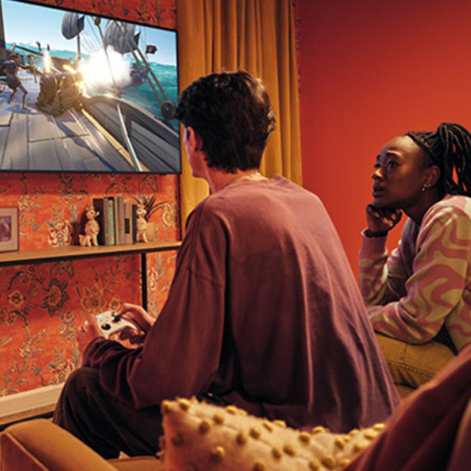 Zwei Personen sitzen in einem Zimmer und schauen auf einen Fernseher, auf dem ein Videospiel gezeigt wird. Ein Mann hält einen Controller.