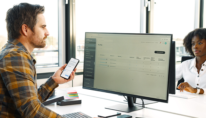 Ein Mann sitzt vor seinem PC und hat ein Smartphone in seiner Hand.