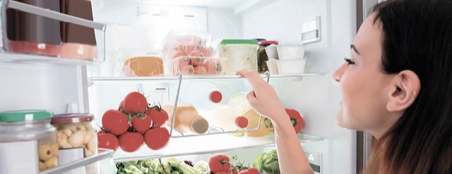 Eine Frau zeigt in einen offenen Kühlschrank mit Regalen voller Lebensmittel wie Tomaten und Behälter.