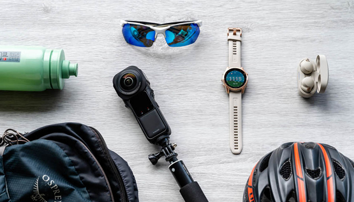 Auf einer hellen Fläche liegen eine blaue Trinkflasche, eine Tasche, eine Kamera, eine Sonnenbrille, eine Smartwatch, Kopfhörer und ein Helm.