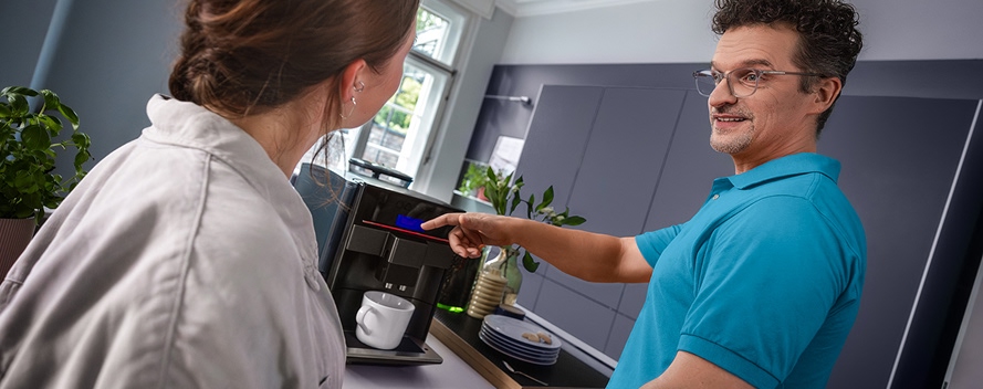 Mann in blauem Poloshirt erklärt einer Frau die Bedienung einer Kaffeemaschine.