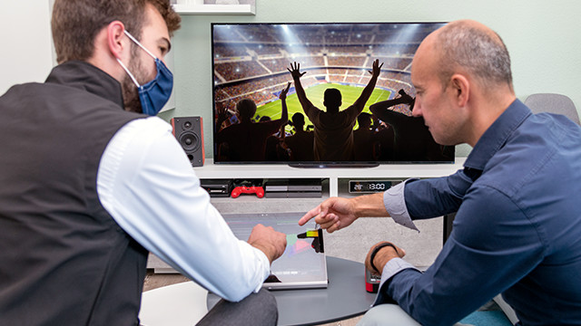 Zwei Männer spielen ein Videospiel vor einem Fernseher, der eine Menschenmenge in einem Stadion zeigt. Einer trägt eine Maske.