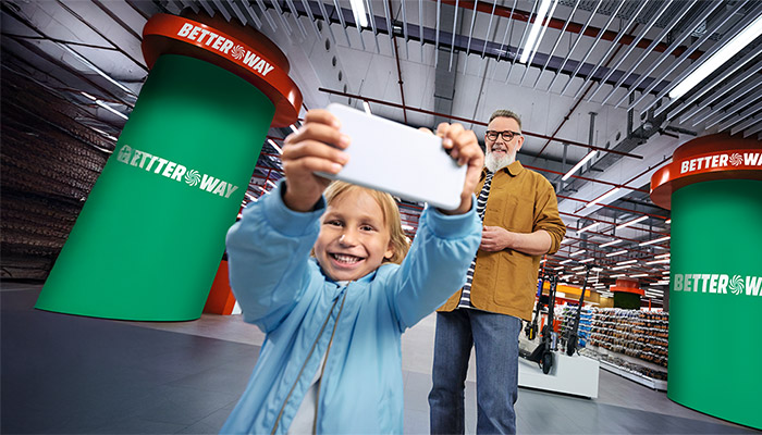 Ein lächelndes Kind hält ein Handy in einem Geschäft, ein Mann steht im Hintergrund, grüne Werbesäulen sind sichtbar.