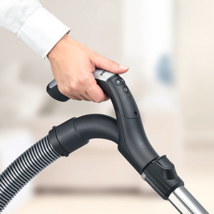 Eine Hand hält einen Staubsaugergriff mit einem grauen Schlauch in einem hellen Raum.