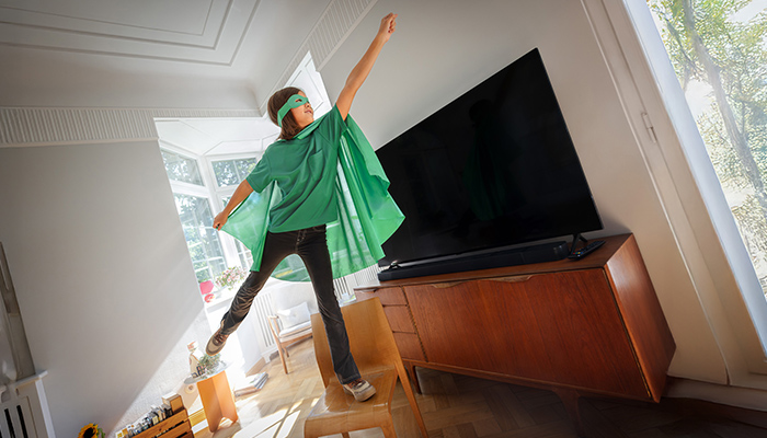 Ein Kind mit grüner Maske und Umhang steht auf einem Stuhl vor einem großen Fernseher in einem hellen Raum.