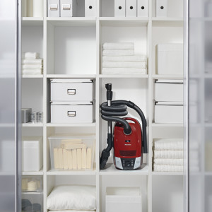 Weißes Regal mit Ordnern, Boxen, Handtüchern und einem roten Staubsauger in der Mitte.