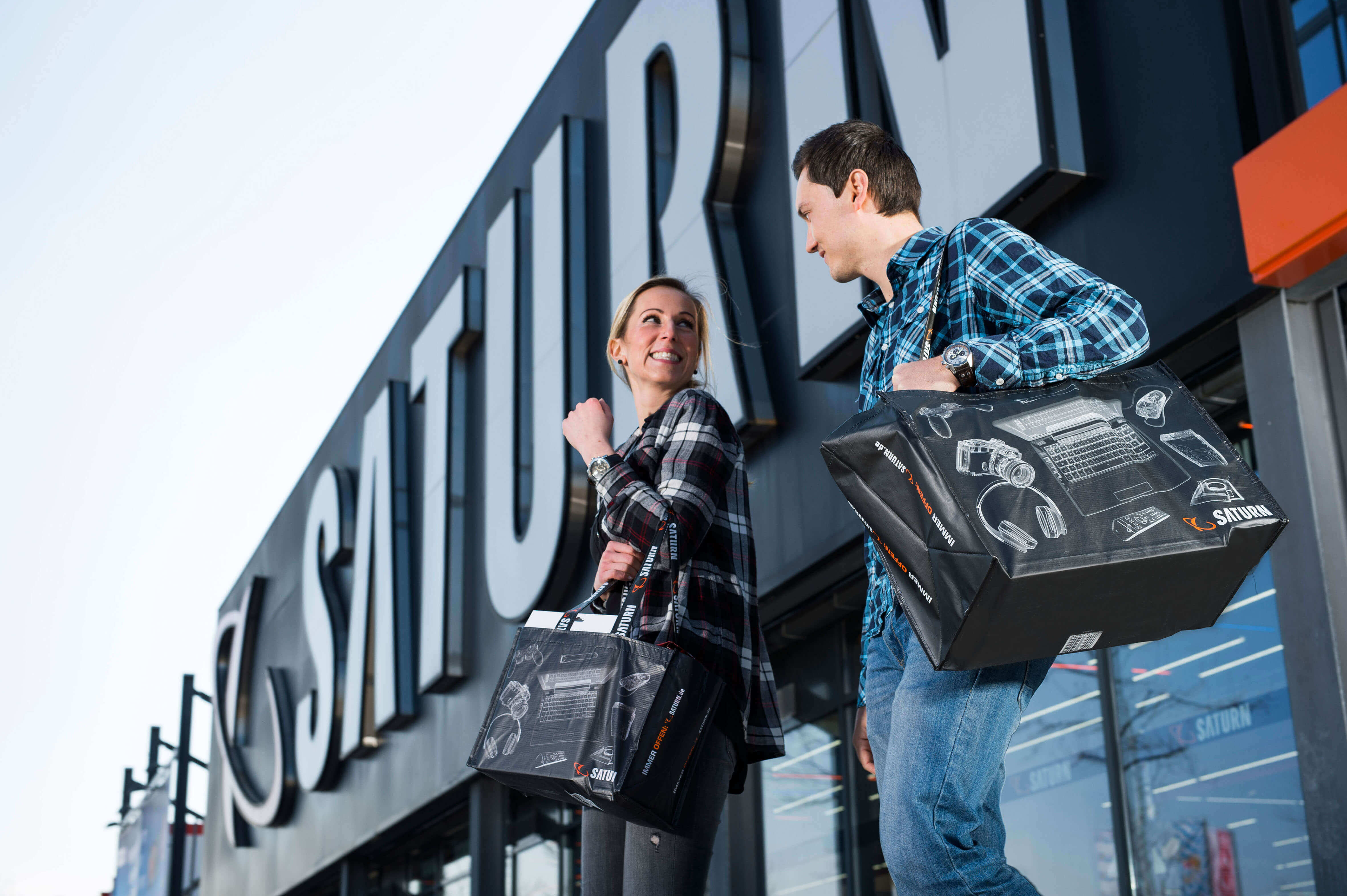 Ein Paar mit Einkaufstüten vor einem Saturn-Geschäft. Die Tüten sind schwarz mit weißen Gerätezeichnungen. Gebäude mit großem Saturn-Schriftzug.