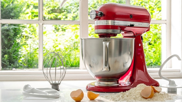 Ein roter Standmixer mit Edelstahlschüssel steht auf einer weißen Arbeitsplatte, daneben Schneebesen, Teigschaber, Mehl und Eierschalen.