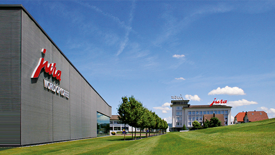 Graues Gebäude mit rotem "Jura"-Logo, gegenüber ein Gebäude mit Turm auf grünem Hügel unter blauem Himmel.