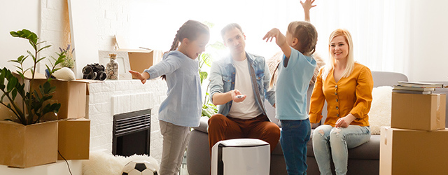 Ein weißer Luftreiniger steht im Vordergrund; im Hintergrund sitzt eine Familie auf einem grauen Sofa.