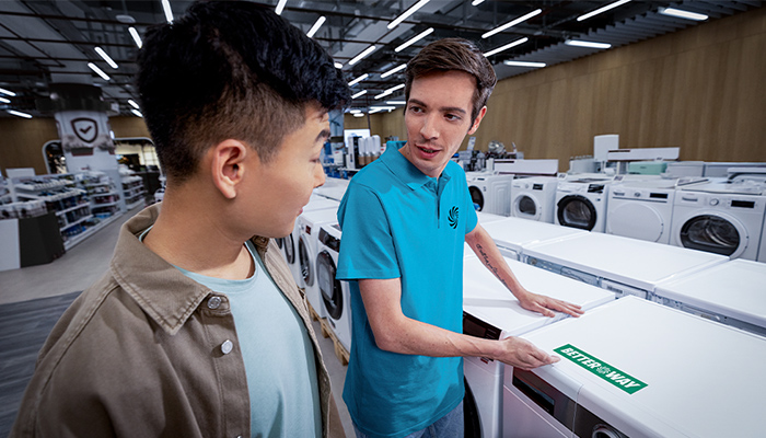 Ein Mann in blauem Hemd zeigt einem anderen Mann in einer Abteilung mit Haushaltsgeräten eine Waschmaschine.