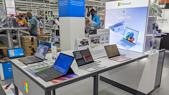 In einem Geschäft stehen mehrere Microsoft-Laptops auf einem Tisch. Im Hintergrund sind Kunden und Regale zu sehen.