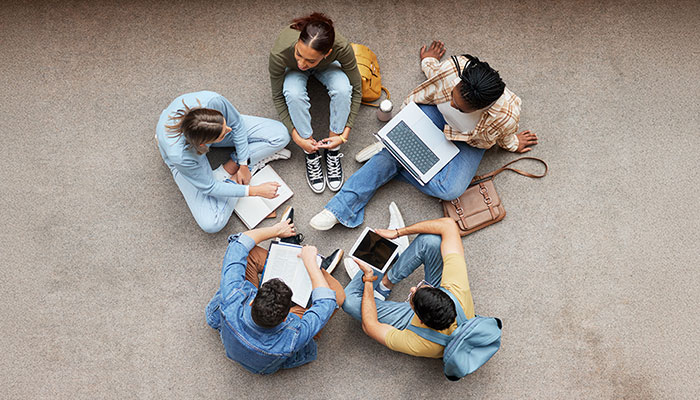 Eine Gruppe von fünf Studierenden sitzt im Kreis auf dem Boden und arbeitet mit Laptops, Tablets und Notizbüchern.
