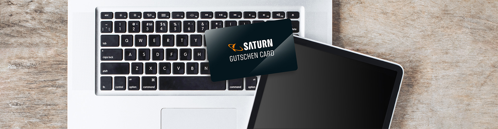 Tastatur eines Laptops mit einer schwarzen Saturn Gutschein-Karte darauf, daneben ein Tablet, auf einem Holztisch.