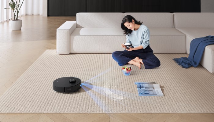 Eine Frau sitzt auf einem Teppich, während ein Roboterstaubsauger in der Nähe reinigt. Ein Sofa und eine Zimmerpflanze sind auch sichtbar.