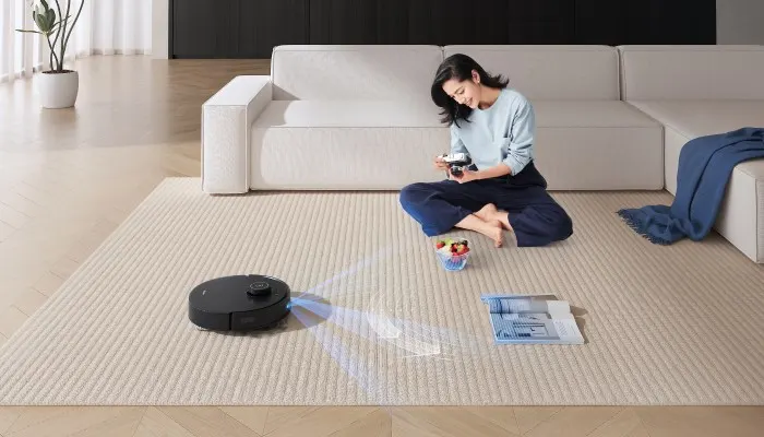 Eine Frau sitzt auf einem Teppich, während ein Roboterstaubsauger in der Nähe reinigt. Ein Sofa und eine Zimmerpflanze sind auch sichtbar.