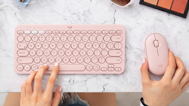 Eine rosa Tastatur und Maus auf einem marmorierten Tisch. Eine Hand tippt, die andere hält die Maus. Kosmetik und Brille sind auch zu sehen.