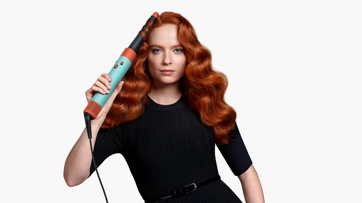 Eine Frau hält ein blau-orangefarbenes Lockenstab vor gewelltem Haar auf einem weißen Hintergrund. Sie trägt ein schwarzes Kleid mit Gürtel.