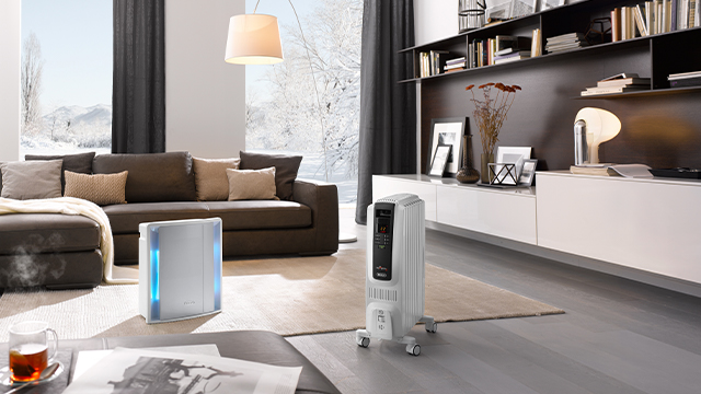 Ein Wohnzimmer mit braunem Sofa, weißem Schrank und Bücherregal. Zwei Heizgeräte stehen auf dem Boden. Draußen ist Schnee zu sehen.