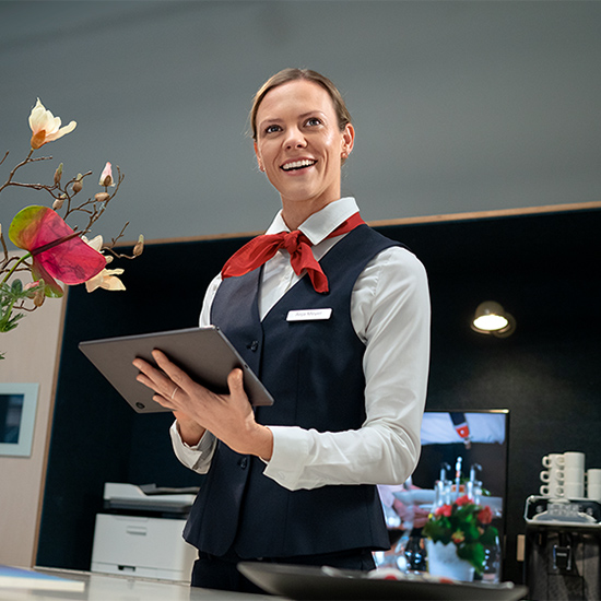 Eine Mitarbeiterin im Service hält ein Tablet. Sie trägt eine rote Schleife und eine Weste. Blumen und Deko stehen auf der Theke.