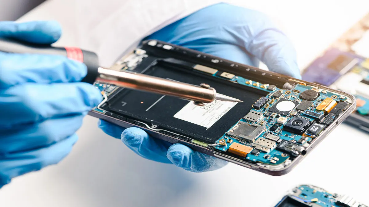 Ein Techniker in blauen Handschuhen repariert ein geöffnetes Smartphone mit einem Lötkolben auf einer weißen Oberfläche.