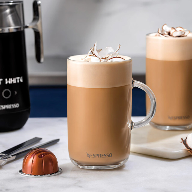 Kaffeegläser mit Milchschaum und Kokoschips stehen auf einer Marmoroberfläche. Eine Kapsel und ein Milchaufschäumer sind auch zu sehen.