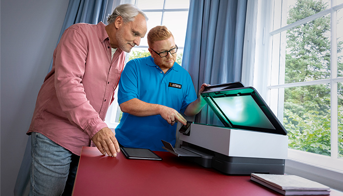 Zwei Personen stehen an einem roten Tisch mit einem Drucker vor einem Fenster mit Vorhängen.