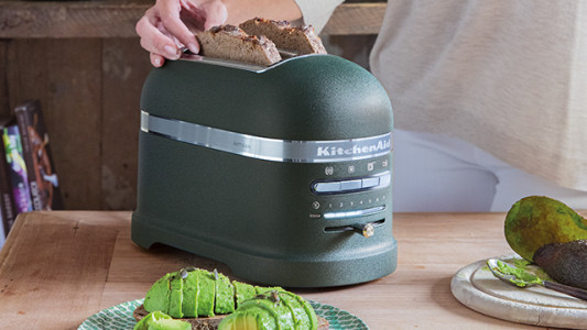 Eine Hand legt Toast in einen grünen Toaster auf einem Holztisch. Auf dem Tisch liegen Teller mit Avocado und Avocados.