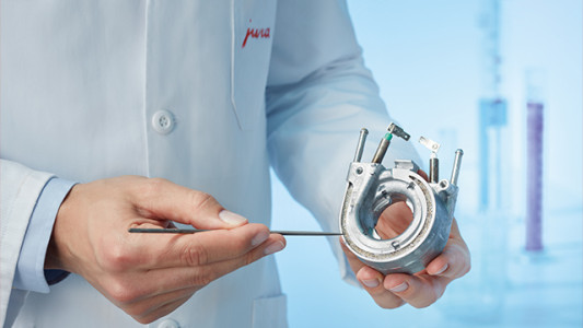 Ein Arzt im weißen Kittel hält ein silbernes medizinisches Gerät mit einer Nadel vor blauem Hintergrund.