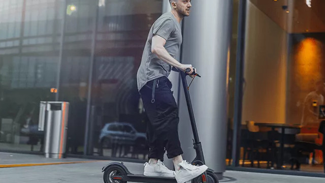 Ein Mann auf einem Elektroroller vor einem Gebäude mit Glasfassade. Im Hintergrund sind ein Mülleimer und ein Auto sichtbar.