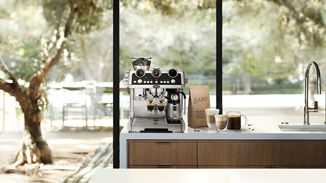 Kaffeemaschine auf einer Küchentheke mit Tassen und einer Kaffeebohne Packung vor einem Fenster mit Blick auf Bäume.