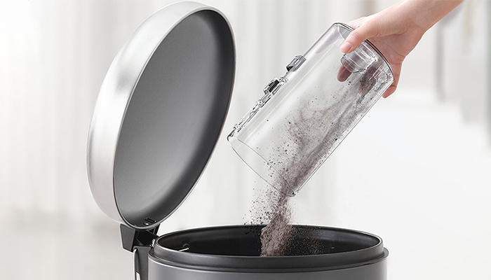 Eine Hand leert einen durchsichtigen Behälter mit Staub in einen geöffneten, grauen Mülleimer mit Deckel.