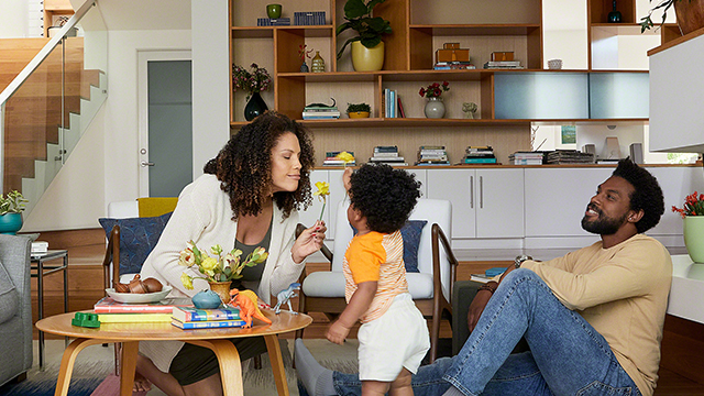 Eine Familie in einem Wohnzimmer: Eine Frau hält einer Person eine gelbe Blume hin, während ein Mann zusieht. Bücherregale im Hintergrund.